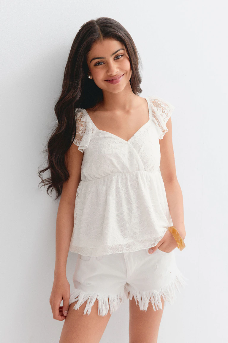 White lace V-neck blouse with flutter cap sleeves and gathered empire waist, paired with white frayed‑hem denim shorts; pearl stud earrings and amber‑tone bangle.