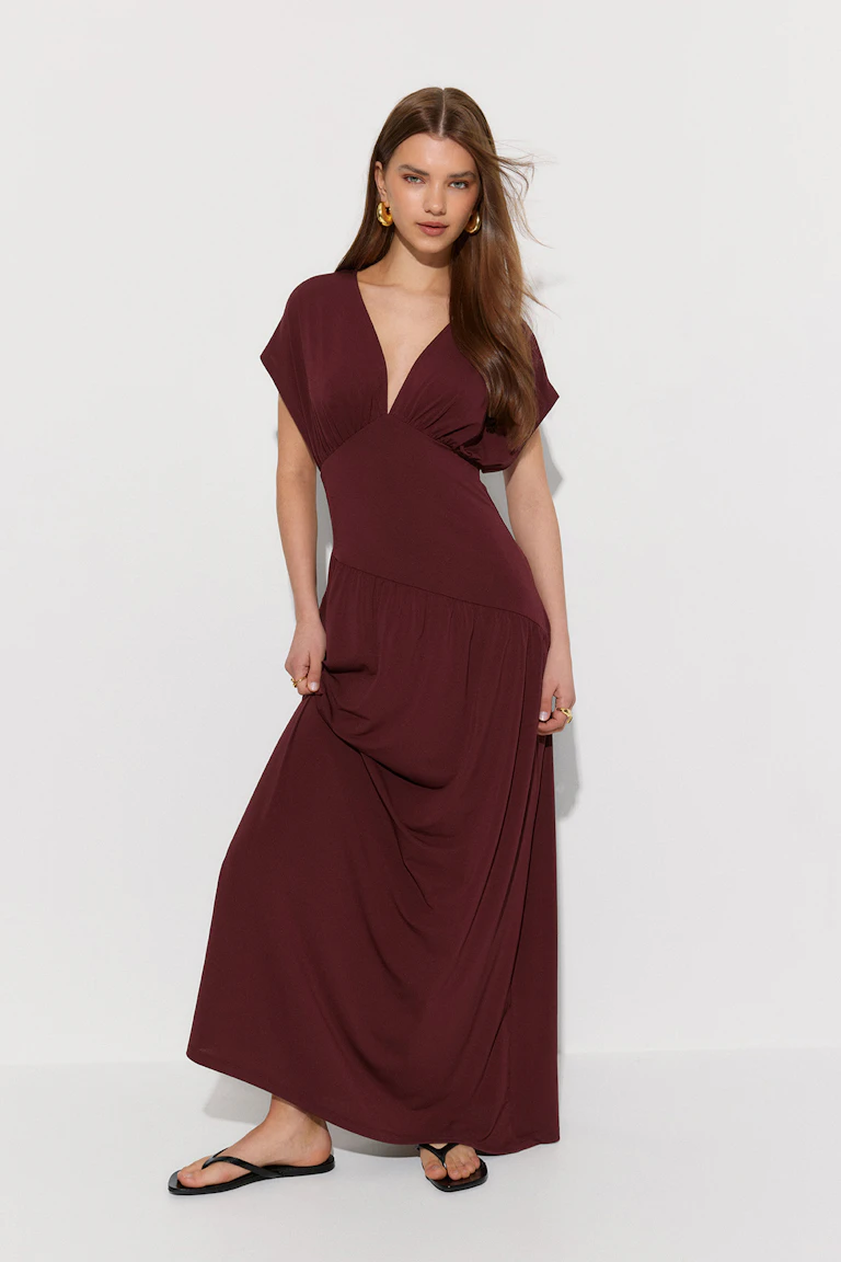 Tief burgunderrotes Maxikleid mit V-Ausschnitt, kurzen Kimonoärmeln und asymmetrischem Saum, kombiniert mit schwarzen Zehenstegsandalen und goldenen Ohrringen und Ringen.