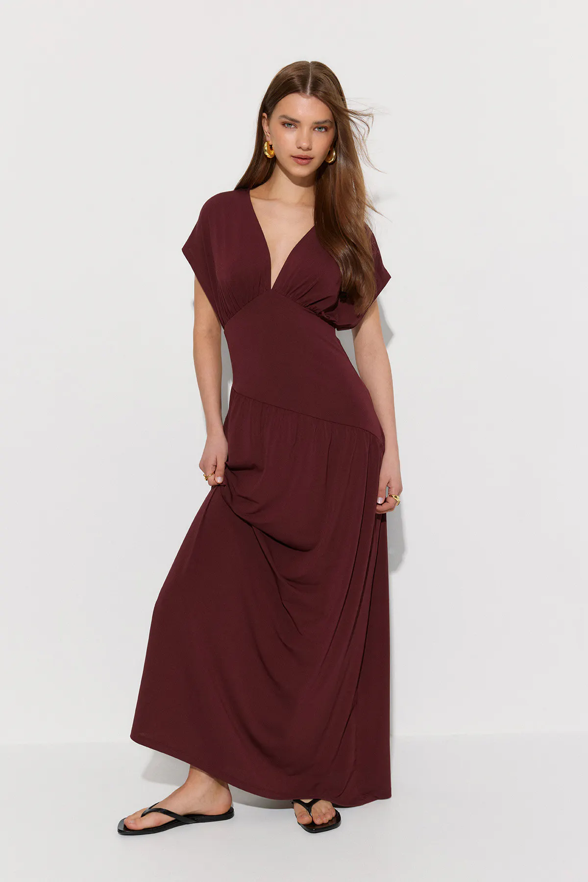 Deep burgundy V-neck maxi dress with short kimono sleeves and an asymmetrical seam, paired with black thong sandals and gold hoop earrings and rings.