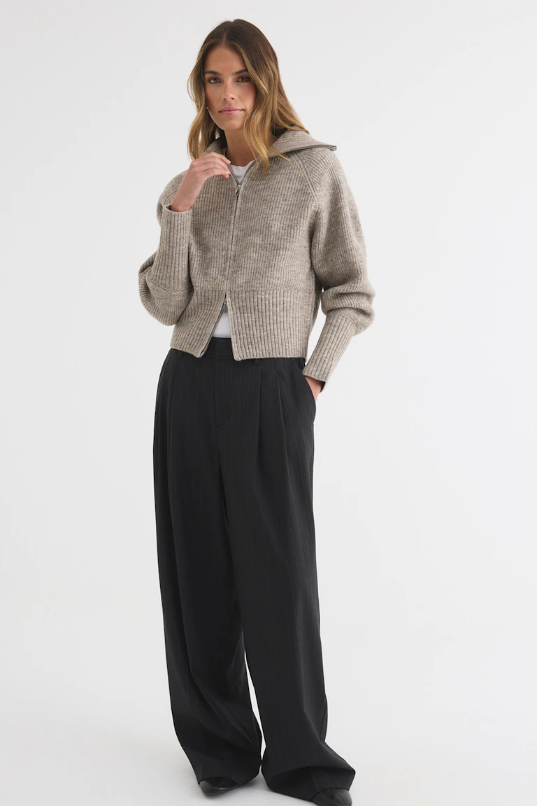 Das Model trägt eine beige, gerippte Strickjacke mit Kapuze und Reißverschluss vorne, ein weißes Oberteil, eine schwarze Hose mit weitem Bein und schwarze Schuhe.