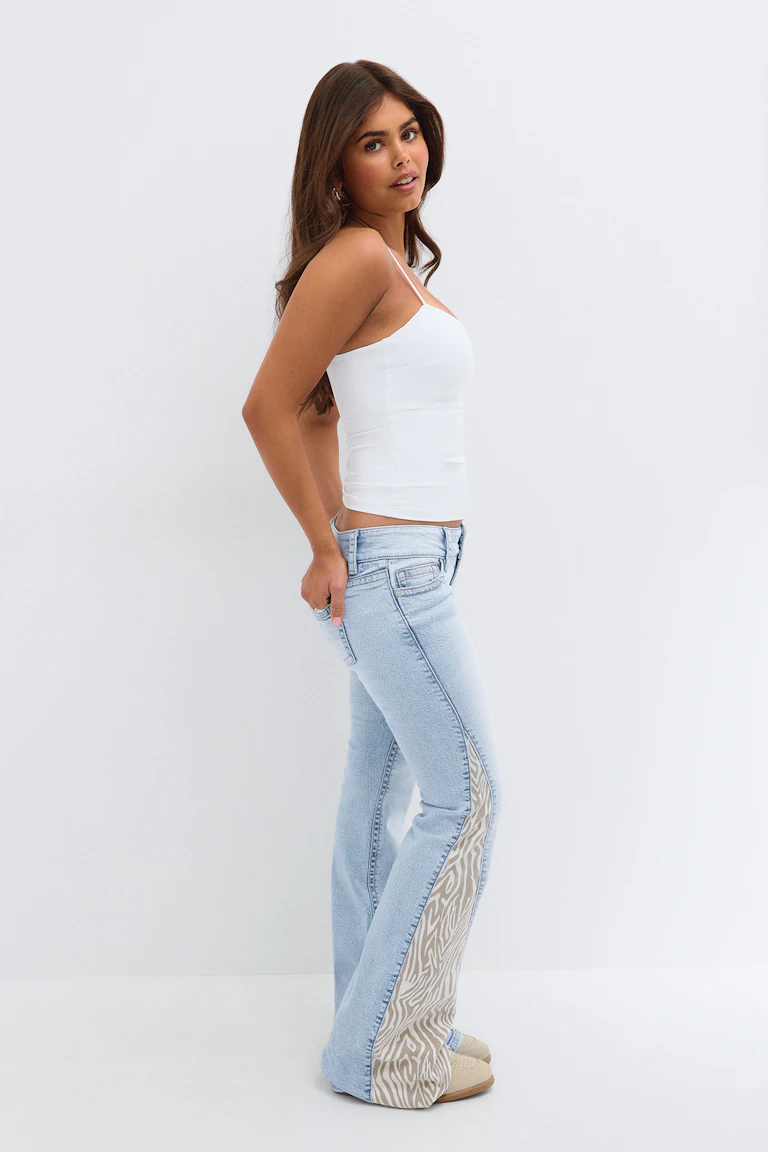 Un haut blanc à fines bretelles, un jean délavé clair orné d'empiècements latéraux à imprimé zèbre et des ballerines beiges.