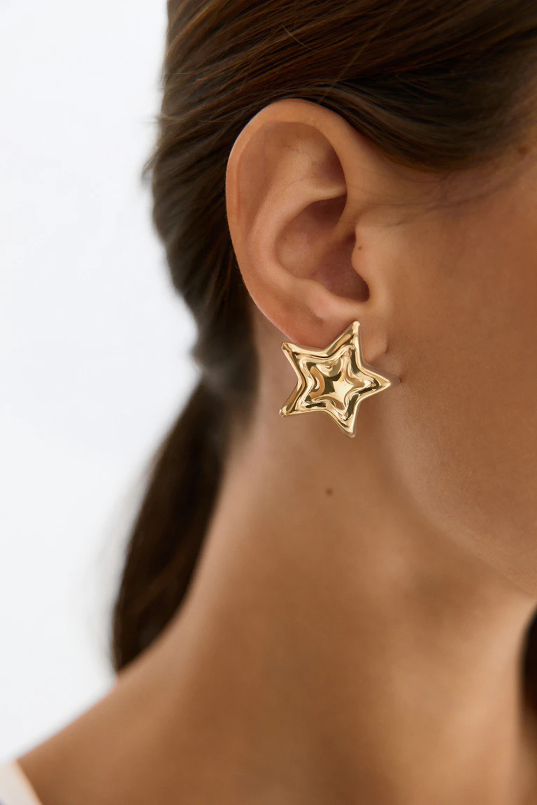 Sie trägt einen polierten, sternförmigen Ohrstecker aus Gold (StyleColorSupplier: 16819).