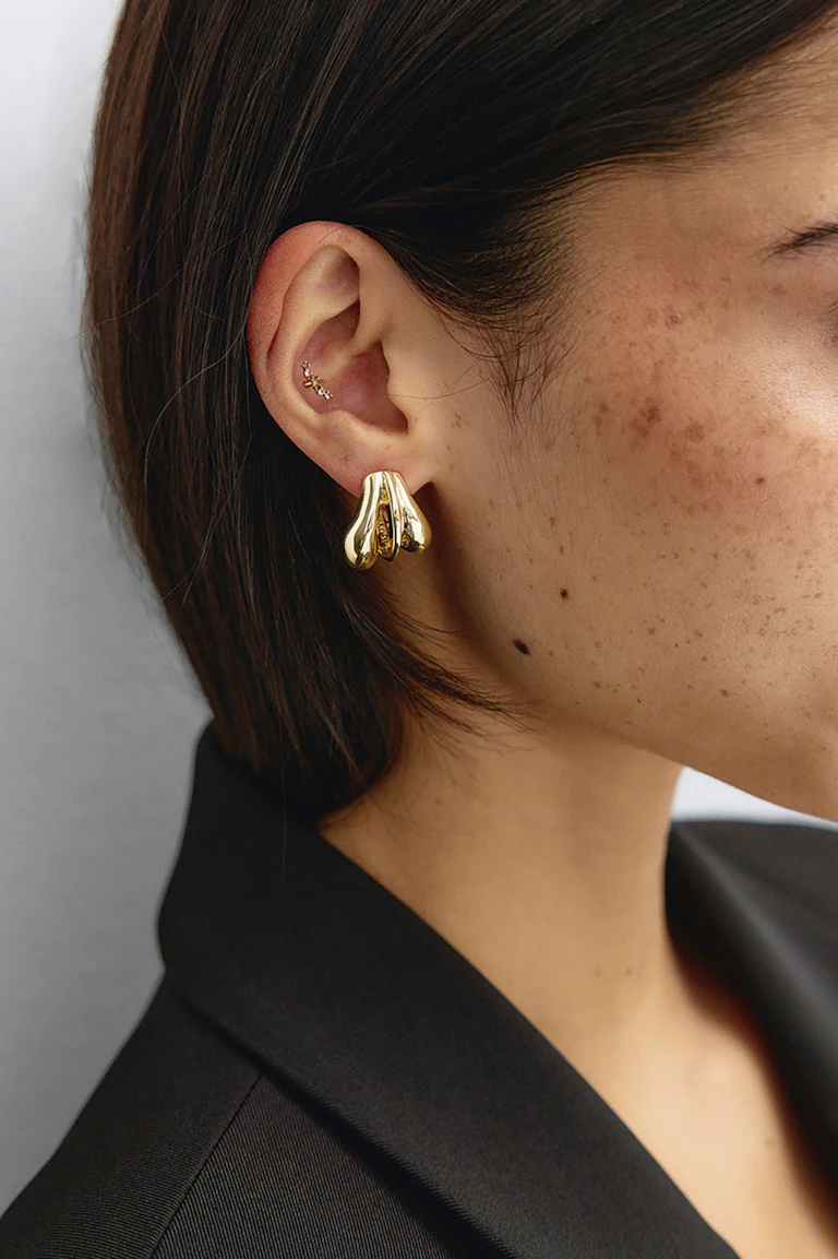 Une boucle d'oreille sculpturale en or poli, en forme de goutte, portée au lobe ; un petit clou en or pour le cartilage, orné de trois pierres transparentes ; et un blazer noir sur mesure.