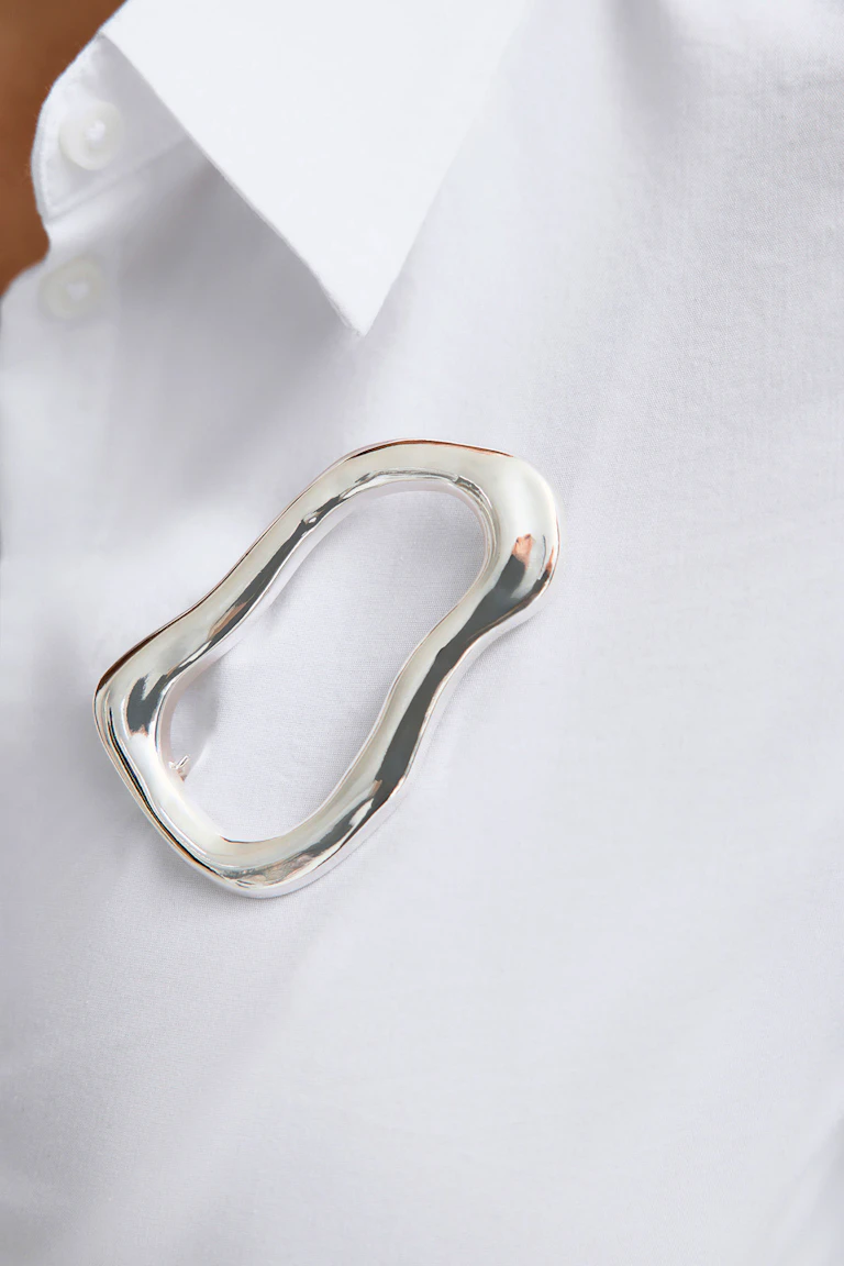 Svart skräddarsydd kavaj med skårad krage, prydd med en oregelbunden, skulptural brosch i silver.