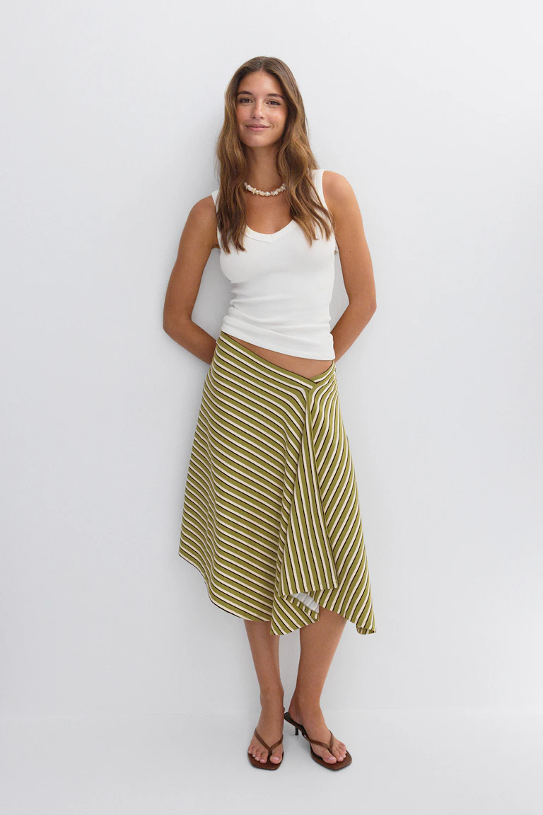 Witte mouwloze V-hals getailleerde tank, olijfgroene en witte diagonaal gestreepte asymmetrische midirok, schelpketting en bruine teenslippers met hak.
