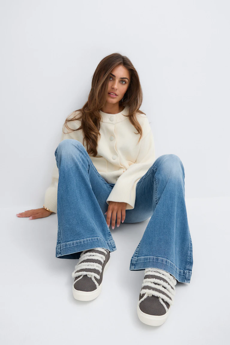 En krämfärgad kofta, blå utsvängda jeans och randiga slip-on-sneakers.
