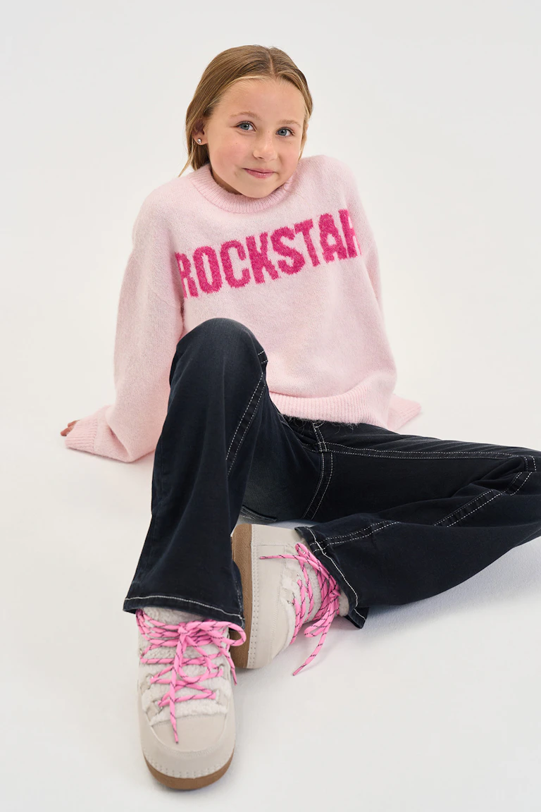 Ein hellrosa, flauschiger Pullover mit magentafarbenem „ROCKSTAR“-Schriftzug, eine dunkle Jeans mit weitem Bein und weiße Sneaker mit rosa Schnürsenkeln.