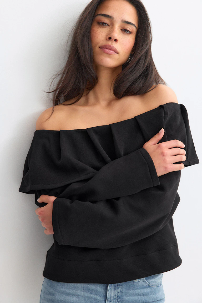 Sort off-the-shoulder top med bred, foldet flæseudskæring og lange ærmer, sat sammen med lysvaskede blå jeans.