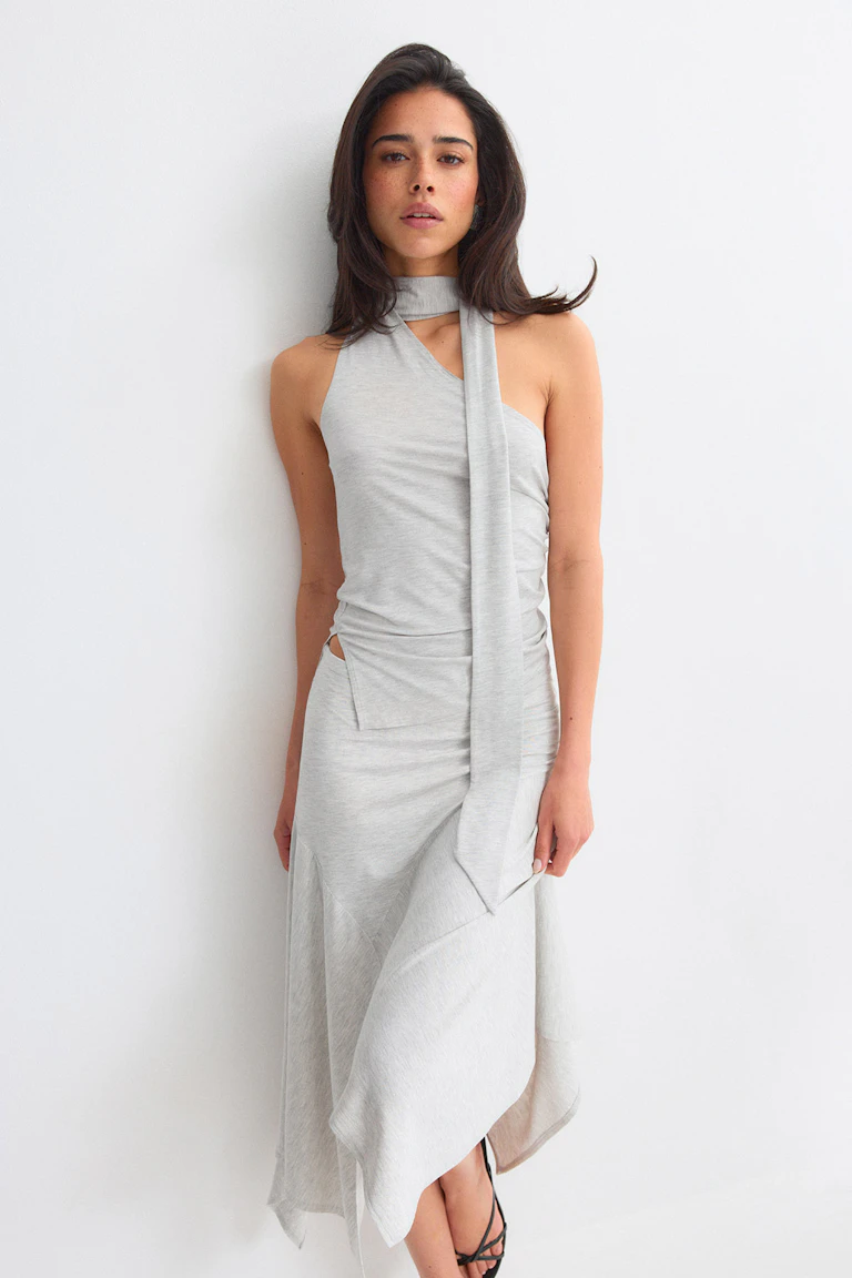 Light gray asymmetrical sleeveless midi dress with halter neck and long scarf tie, ruched side with a small hip cutout and a draped layered skirt, paired with black strappy sandals.