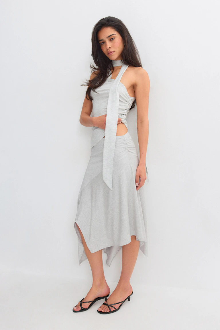 Lichtgrijze mouwloze haltertop met lange sjaal en uitsnede in de taille aan de zijkant, gecombineerd met een bijpassende asymmetrische midirok met zakdoekzoom; zwarte sandalen met lage hakken en bandjes.