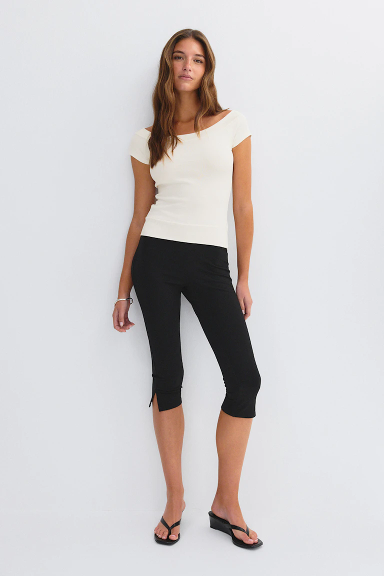Off-white getailleerde top met korte mouwen en boothals, gecombineerd met een zwarte slim-fit capribroek met kleine splitjes in de zoom aan de voorkant, zwarte teenslippers met sleehak en een zilveren armband.