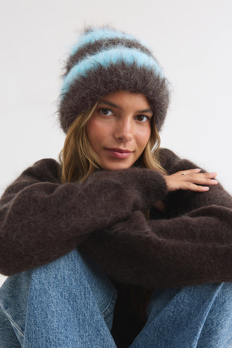 Model iført en mørkebrun, lodden sweater, en blå og brun strikket hue og blå jeans.
