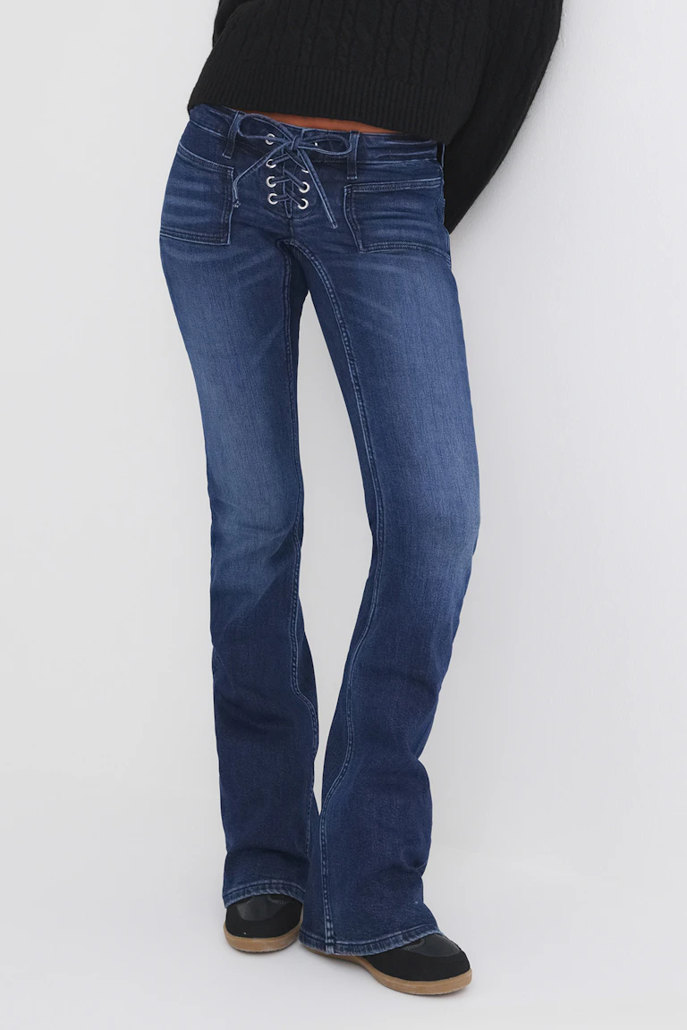 Dunkelblaue, ausgestellt geschnittene Jeans mit Schnürung und aufgesetzten Taschen vorne, kombiniert mit einem schwarzen, gerippten Pullover und schwarzen Schuhen.