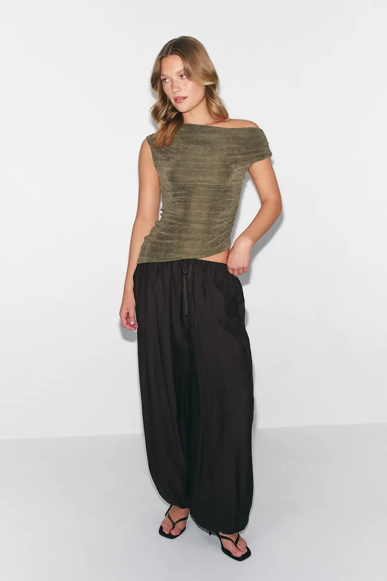 Olijfgroene off-the-shoulder top met korte mouwen gecombineerd met een losse zwarte broek met wijde pijpen en zwarte sandalen met bandjes.