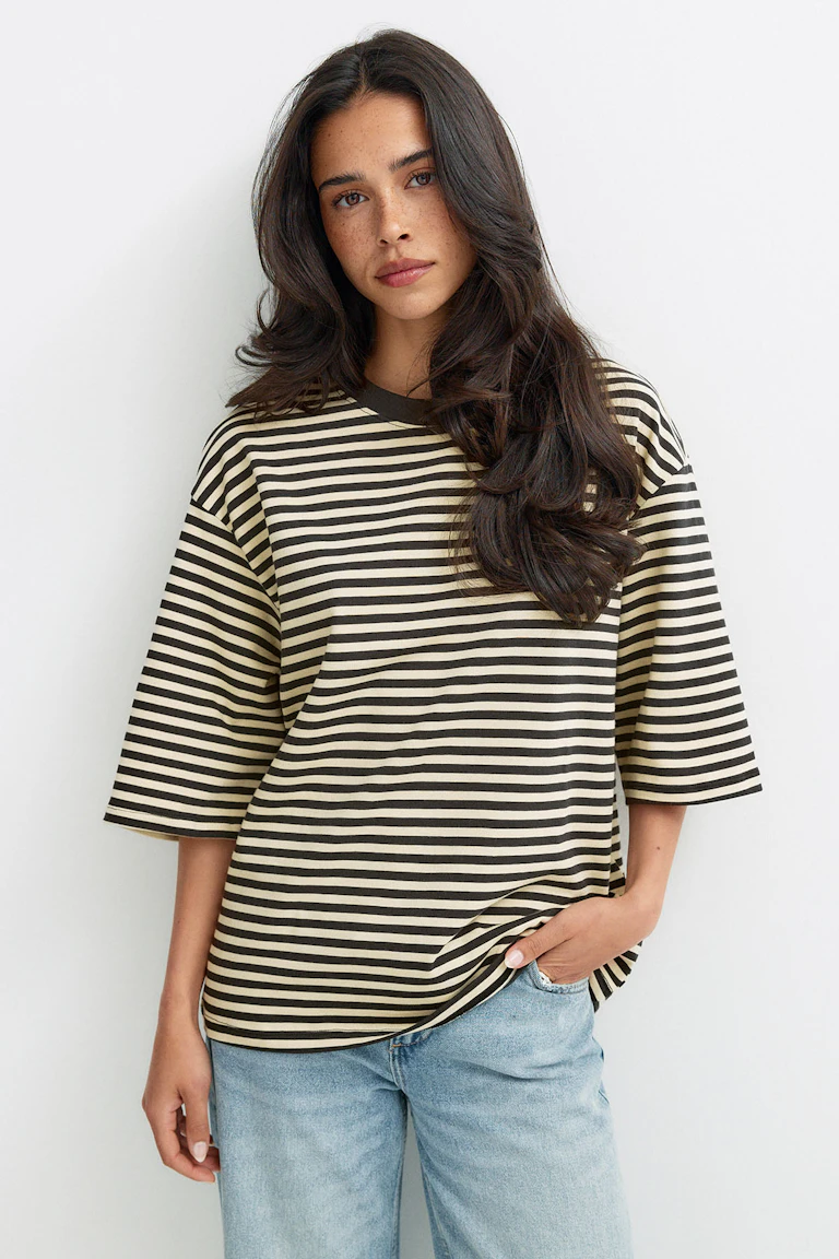 Oversized T-shirt met crèmekleurige en zwarte horizontale strepen, een donkere geribde ronde hals en mouwen tot aan de elleboog, gecombineerd met een lichtgekleurde jeans met rechte pijpen.