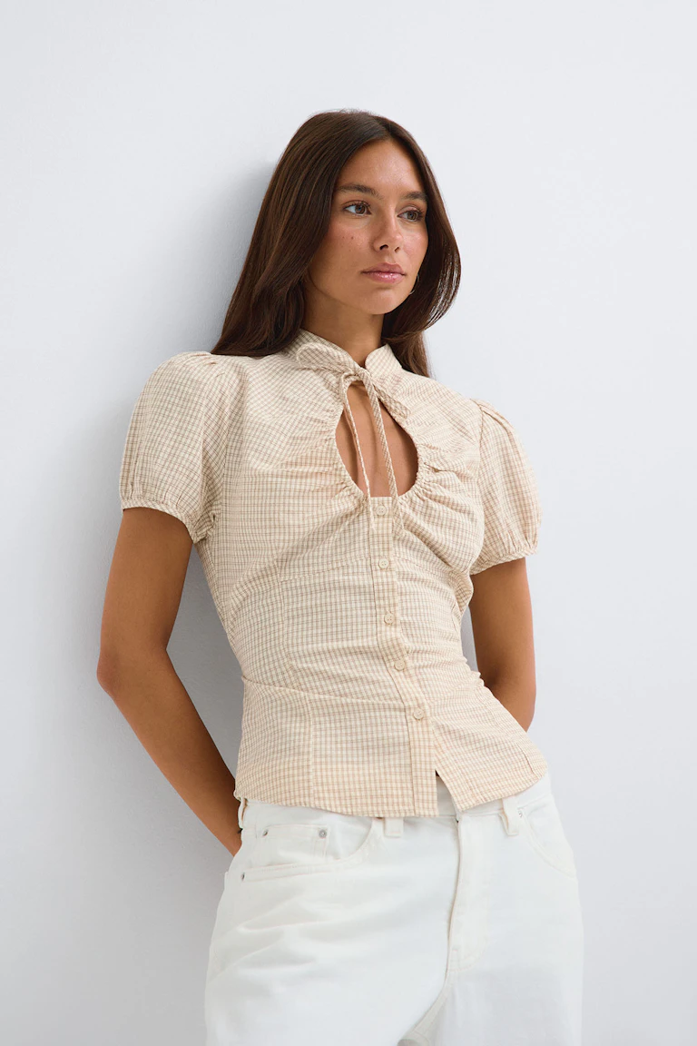 Crèmekleurige geruite blouse met korte mouwen, pofmouwtjes, een strik in de hals en een witte broek.