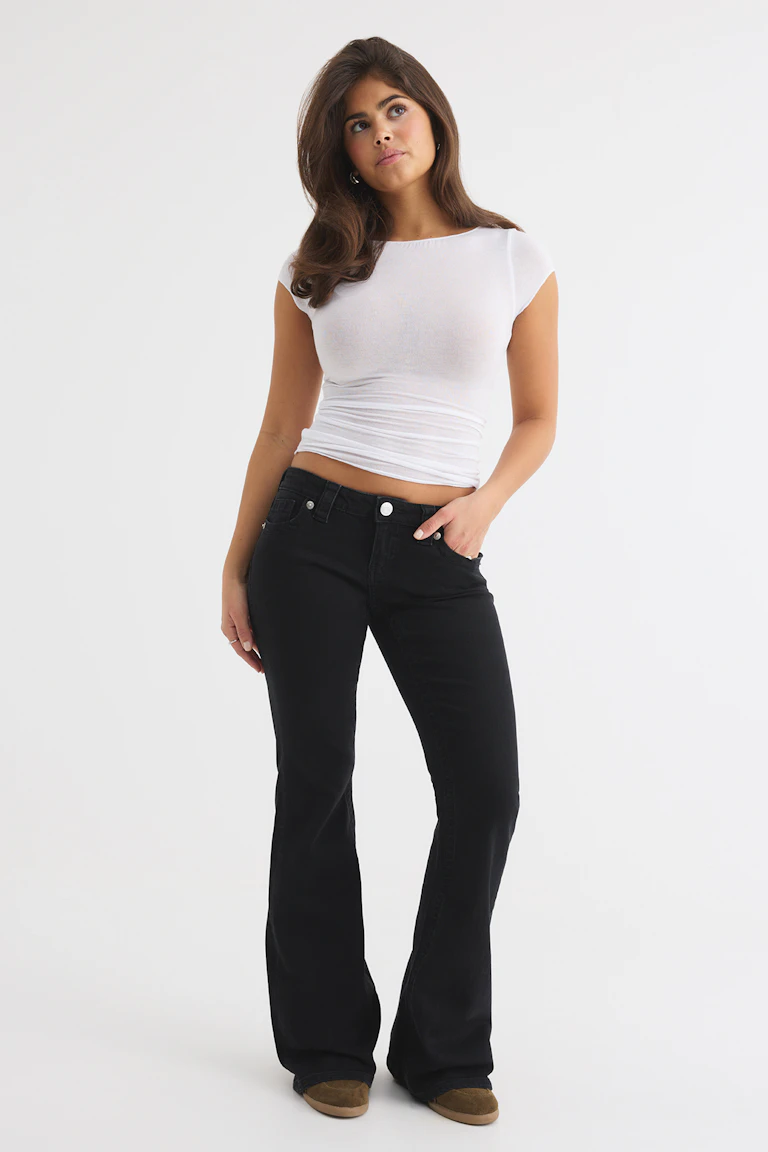 Weißes, figurbetontes Kurzarm-Oberteil, schwarze Schlagjeans und braune Schuhe.