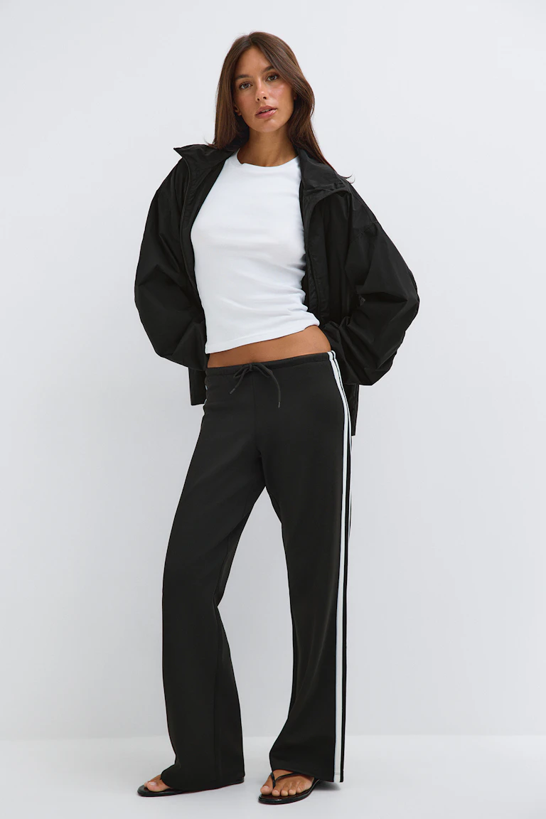 ID 341136, StyleColorSupplier 11519-E: Weißes, figurbetontes T-Shirt, schwarze Jacke mit Reißverschluss, schwarze Hose mit weitem Bein und weißen Seitenstreifen sowie schwarze Sandalen.