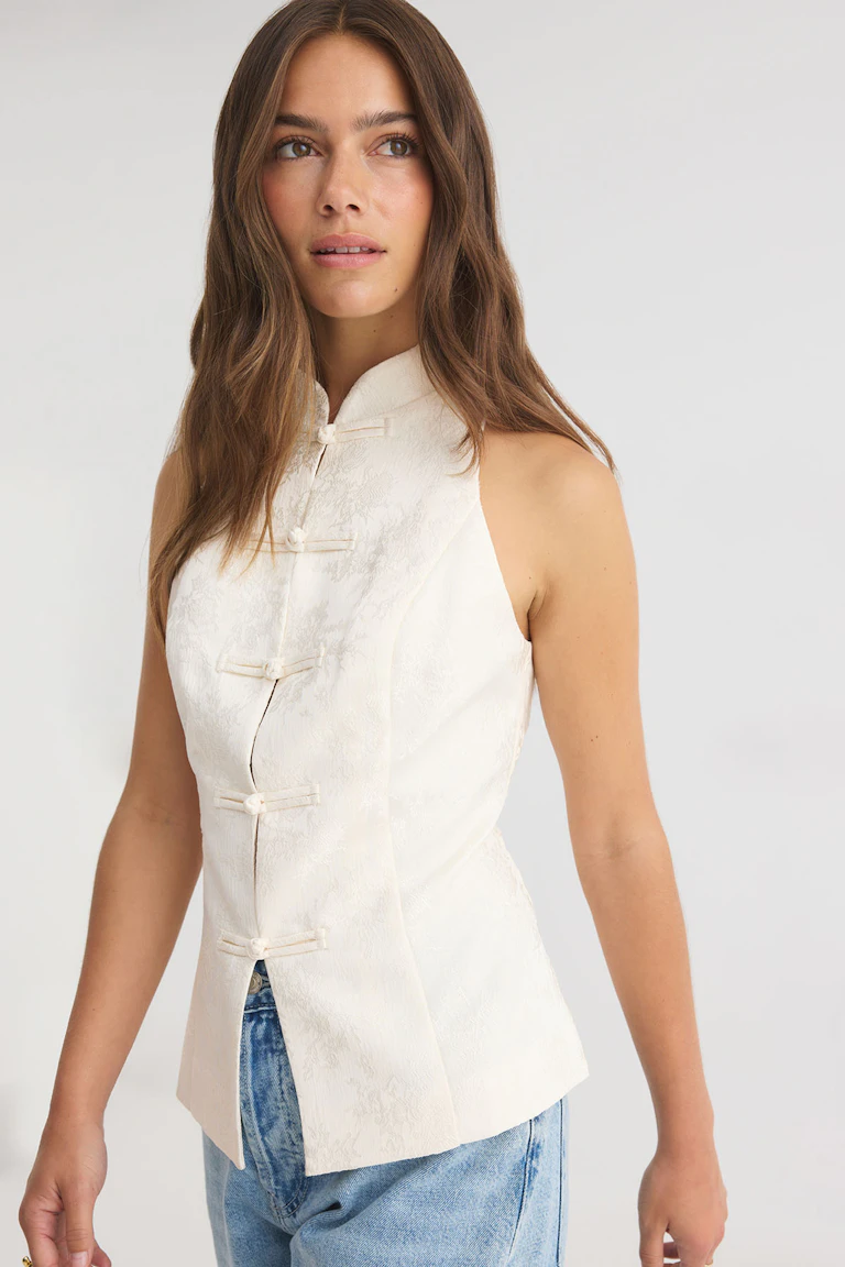 Cream sleeveless mandarin-collar top with frog closures, paired with blue jeans.