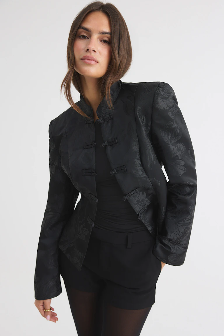 Black floral jacquard jacket with frog closures, worn over a black top, tailored black shorts, and sheer tights.