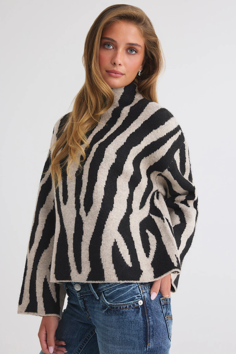 Ylisuuri musta-beige zebrakuvioinen neulottu turtleneck-villapaita, yhdistettynä sinisiin farkkuihin.