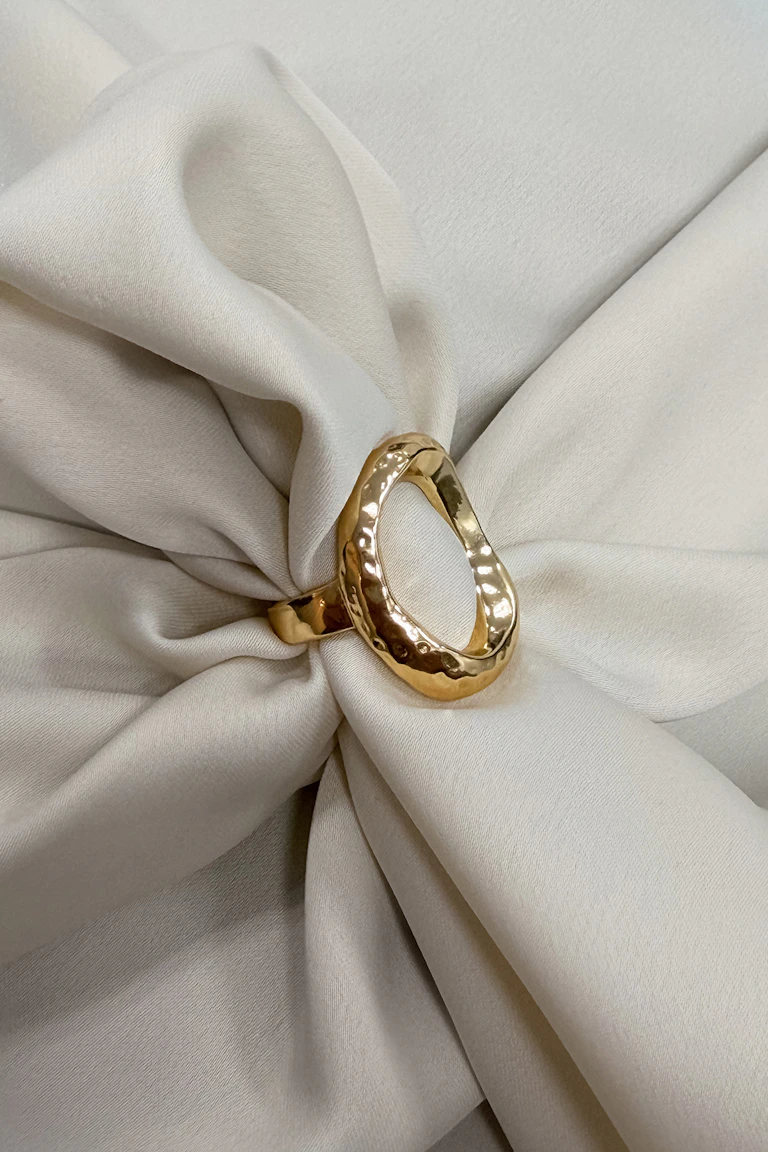Ovaler Ring aus gehämmertem Gold, präsentiert auf elfenbeinfarbenem Stoff.