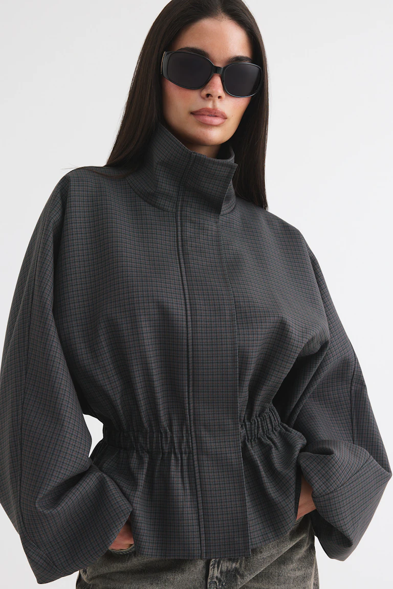 Ein Model trägt eine übergroße schwarze Sonnenbrille und eine dunkle, karierte Jacke mit hohem Kragen und voluminösen Ärmeln.