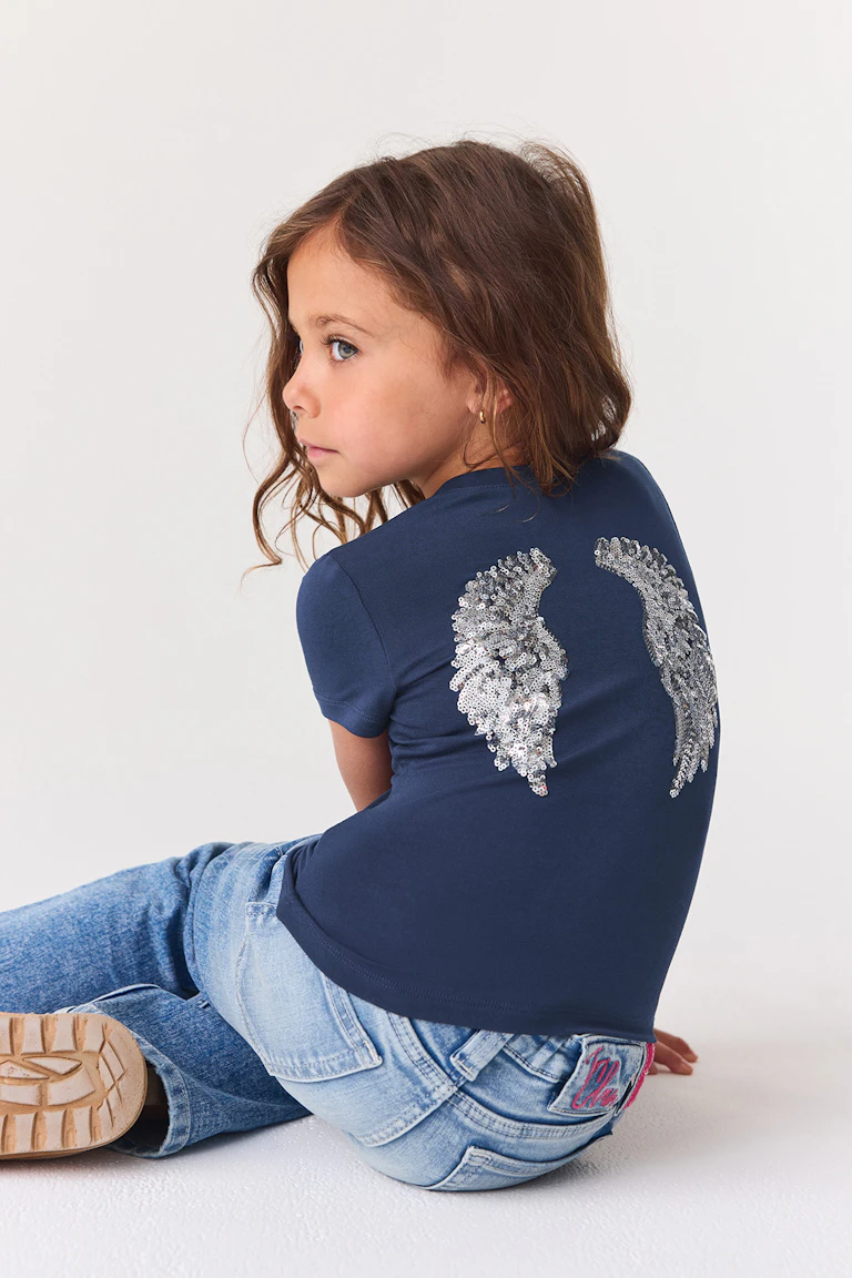 ID 342587, StyleColorSupplier 11519-E: Marineblå T-skjorte med korte ermer, med påsydde vinger med sølvpaljetter og jeans i lys vask.