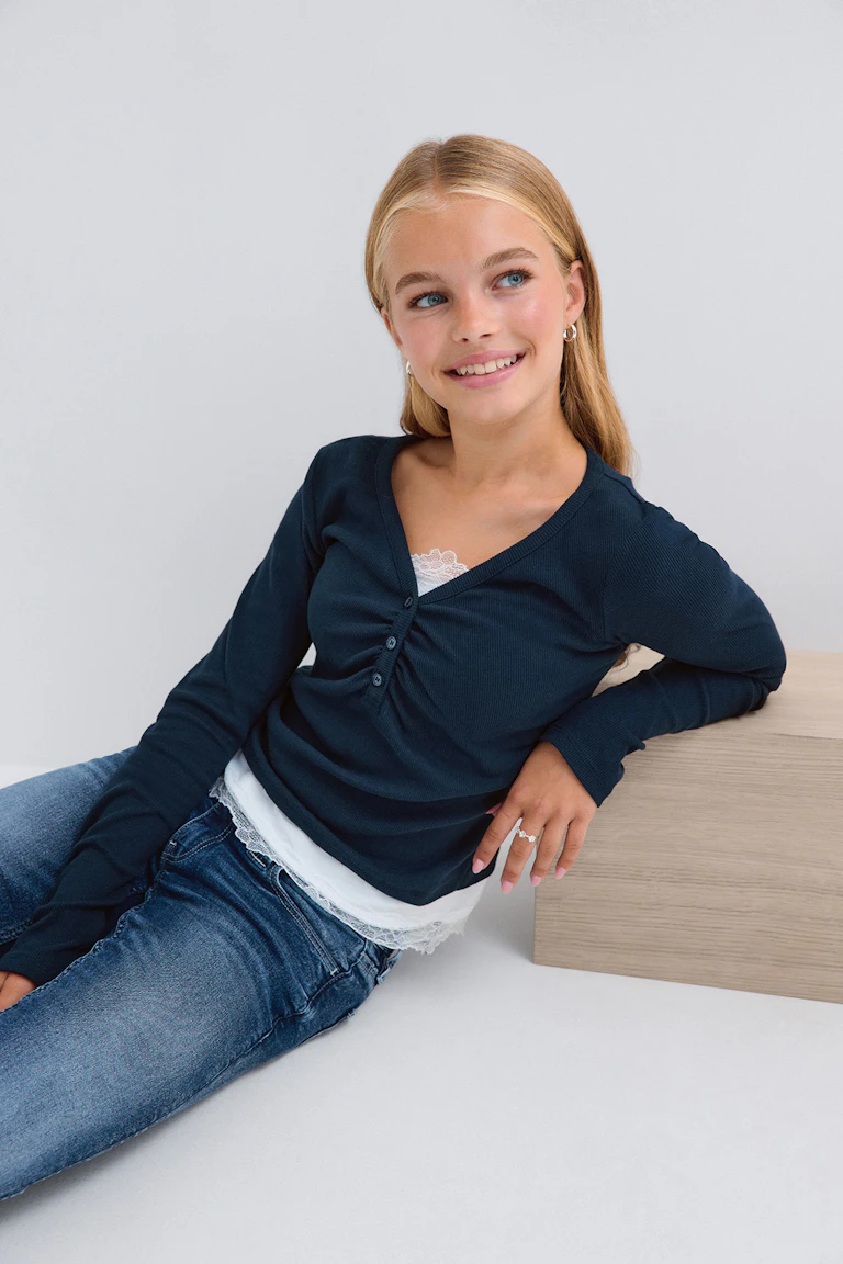 ID 344709, StyleColorSupplier 11519-E: Marineblaues Langarm-Oberteil mit Knopfleiste über einem weißen, mit Spitze besetzten Trägertop, dazu blaue Jeans.