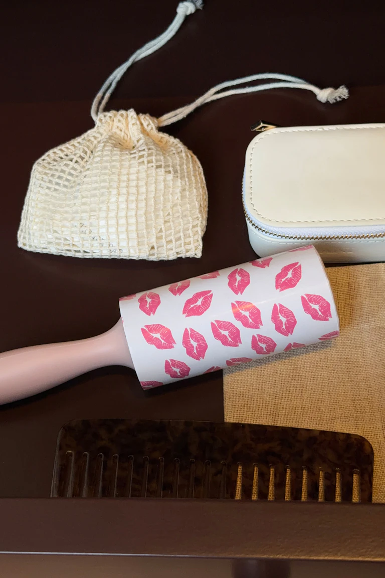 Cremefarbener Beutel aus Netzstoff mit Kordelzug, weißes Etui mit Reißverschluss, rosa Fusselroller mit Lippenabdruck, Kamm in Schildpattoptik und hellbrauner gewebter Beutel.