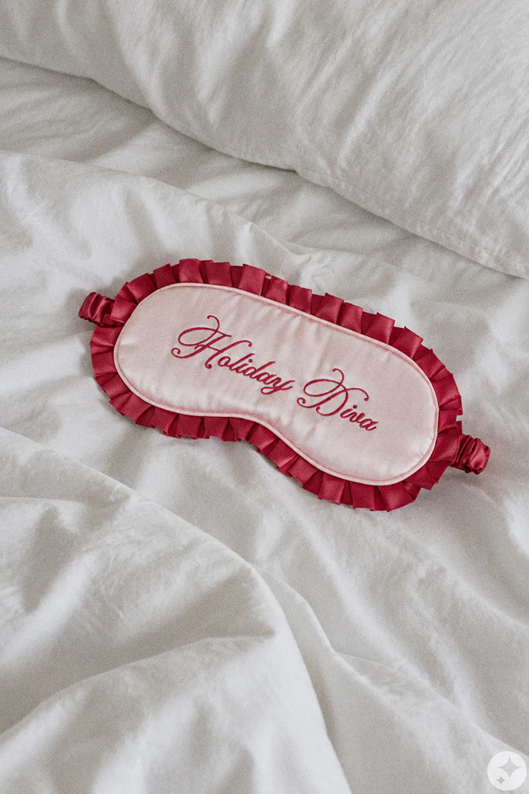 Pink satin eye mask with burgundy ruffled trim and embroidered “Holiday Diva” text.