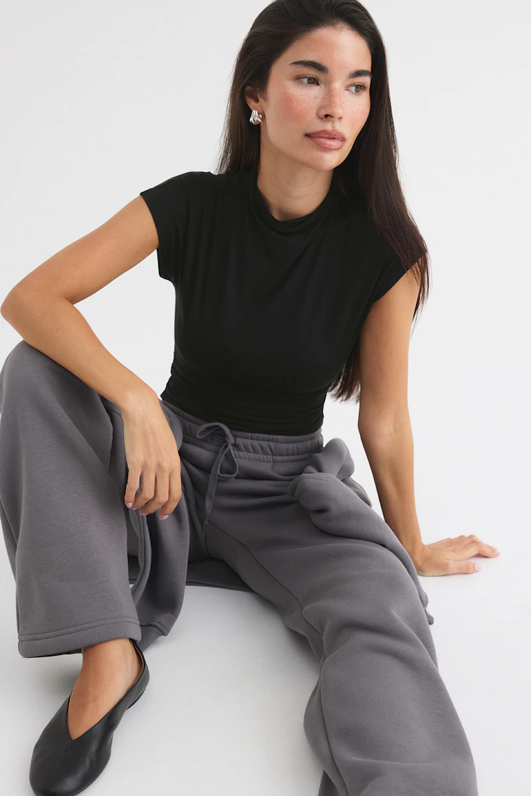 Schwarzes, ärmelloses Oberteil, graue Jogginghose mit weitem Bein und schwarze Ballerinas.
