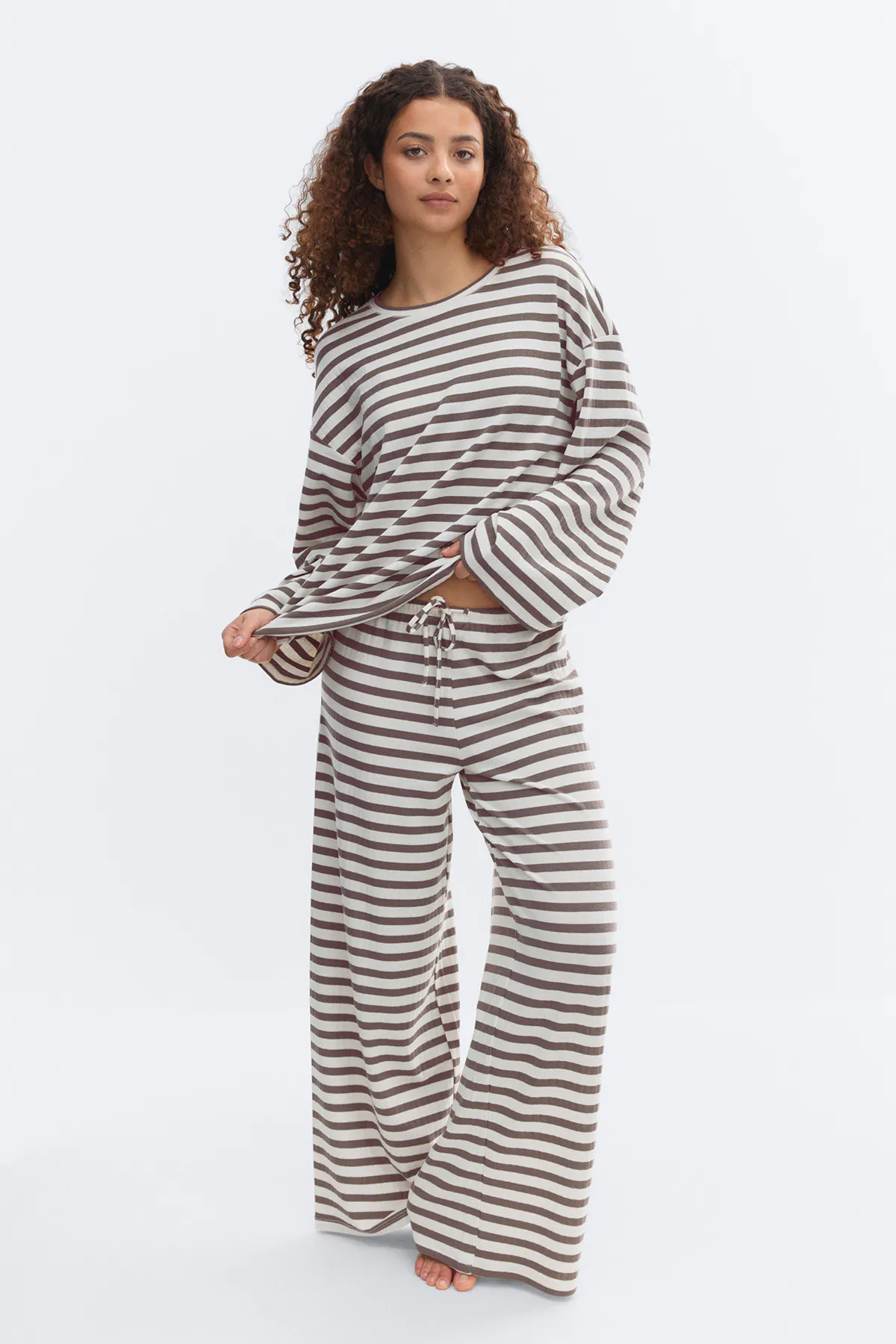 Two-piece loungewear set: long-sleeve crewneck top with wide sleeves and matching wide-leg drawstring pants in horizontal cream and brown stripes; model barefoot.